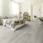 Stylish apartment interior with white walls and herringbone wooden floor. A view from a bedroom with a big bed to another room with an armchair. Real photo.